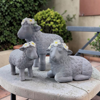 Drei graue Deko-Schafe aus Resin mit weißen Blumenkränzen stehen und sitzen gemeinsam auf einer Steinplatte vor unscharfem Gartenhintergrund.