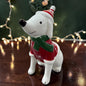 Sitzender weißer Tiziano Keramikhund mit rotem Pullover, grünem Schal und rot-weißer Weihnachtsmütze vor dunklem Hintergrund mit Lichterkette.