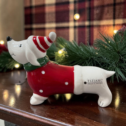 Liegender weißer Tiziano Keramikhund mit rotem Pullover, grünem Schal und rot-weißer Weihnachtsmütze vor weihnachtlich dekoriertem Hintergrund.
