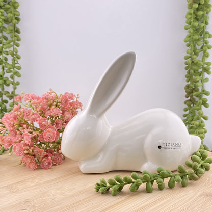 Ein schlafender Keramik Osterhase, der auf dem Boden liegt und seine Ohren ausstreckt. Der Dekohase ist aus creme-weißer Keramik gefertigt, 19cm hoch und im Ambiente abgebildet.