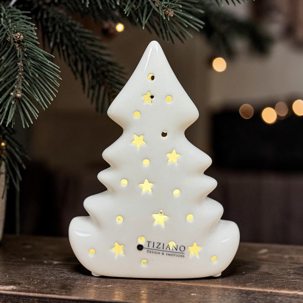 Cremeweißer Tiziano Keramik Baum Alberi mit warm beleuchtetem Stern- und Lochmuster vor Tannenzweigen und Lichtern.