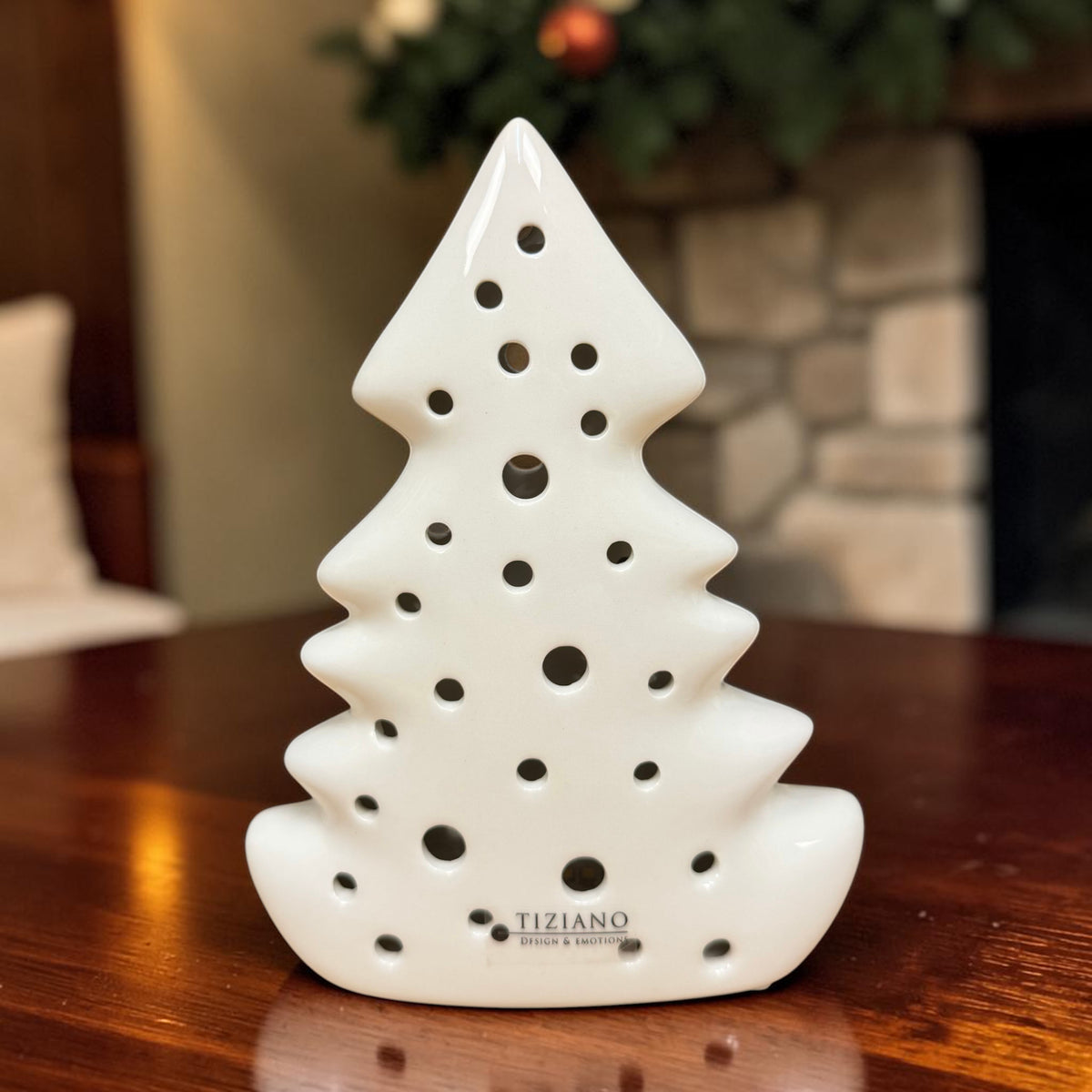 Cremeweißer Tiziano Keramik Baum Alberi mit rundem Lochmuster unbeleuchtet auf einem Holztisch vor weihnachtlichem Hintergrund.