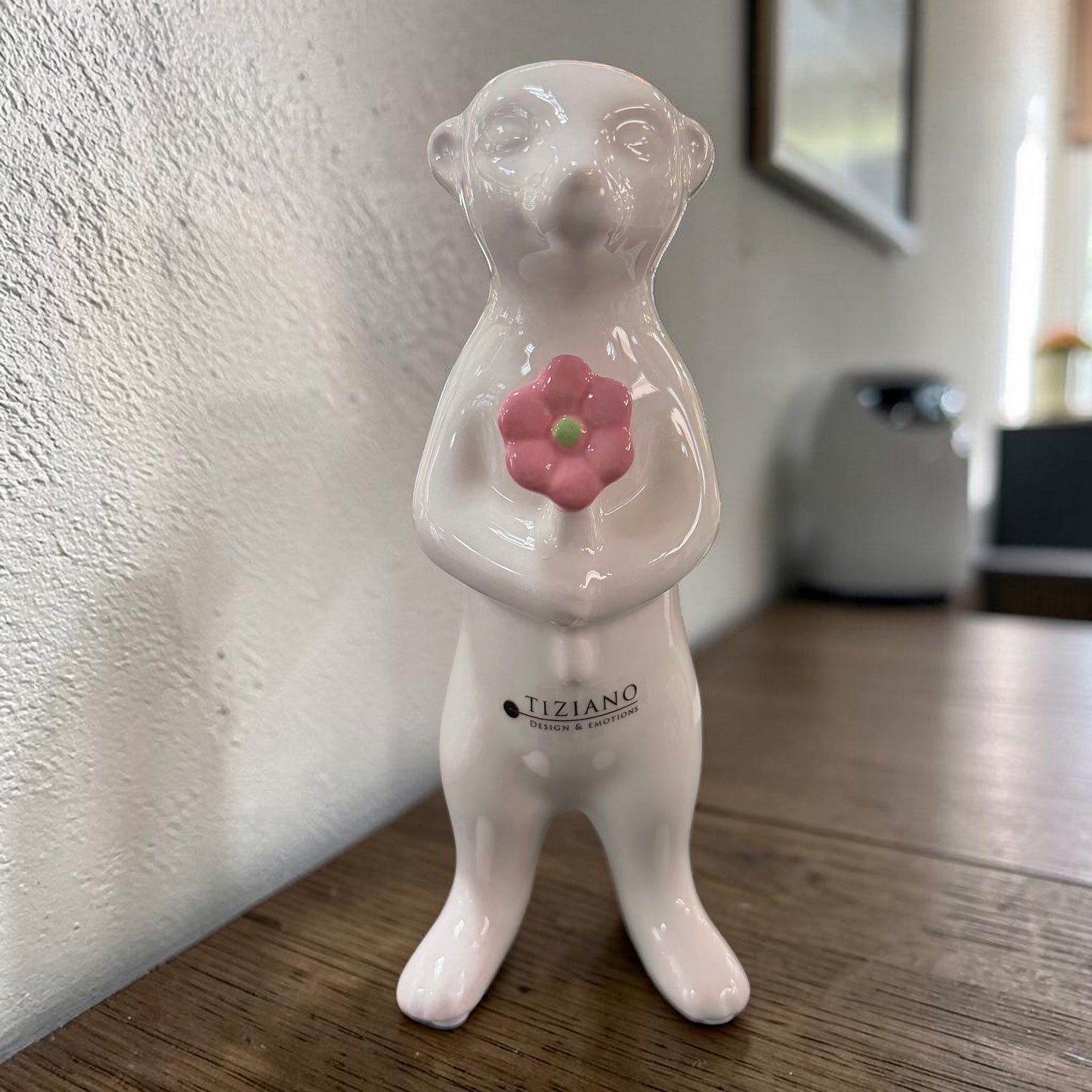 Ein weißes Keramik-Erdmännchen mit rosa Blume steht auf einer Holzoberfläche vor einer hellen Wand und unscharfem Hintergrund.
