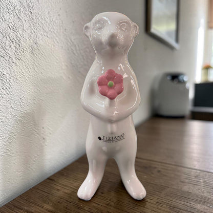 Ein weißes Keramik-Erdmännchen mit rosa Blume steht auf einer Holzoberfläche vor einer hellen Wand und unscharfem Hintergrund.