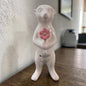 Ein weißes Keramik-Erdmännchen mit rosa Blume steht auf einer Holzoberfläche vor einer hellen Wand und unscharfem Hintergrund.
