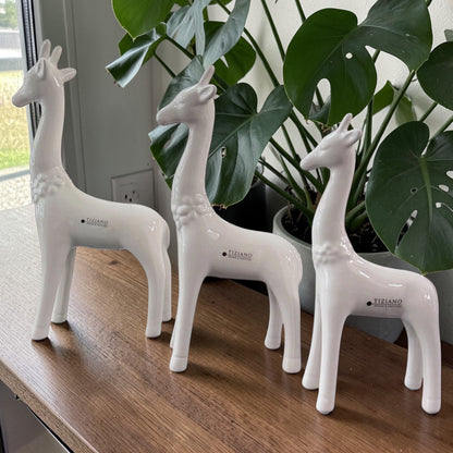 Drei weiße Keramik-Giraffen in verschiedenen Größen stehen dekorativ auf einem Holztisch vor einer grünen Zimmerpflanze.