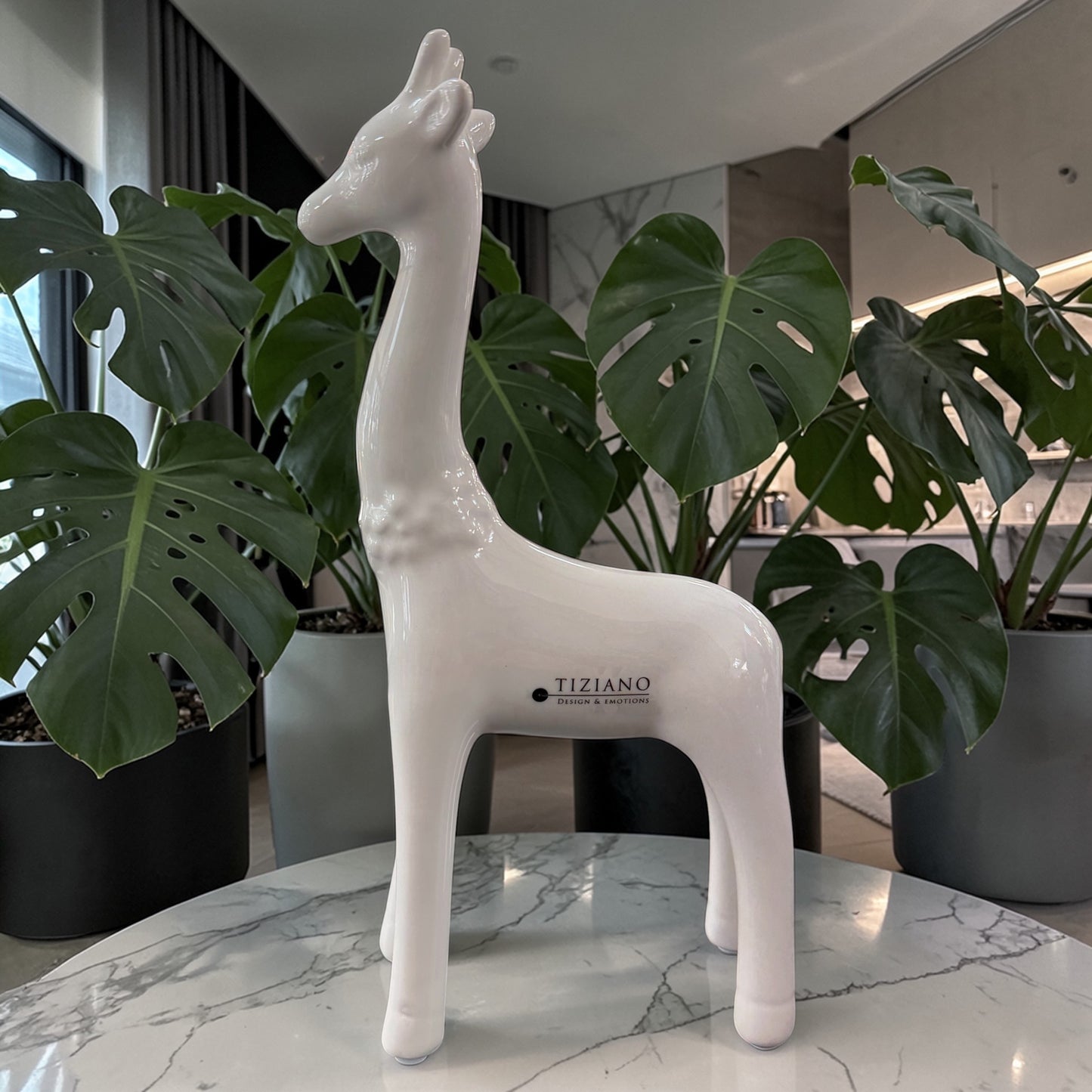 Eine große weiße Keramik-Giraffe steht dekorativ auf einem Marmortisch vor grünen Zimmerpflanzen in modernem Wohnambiente.