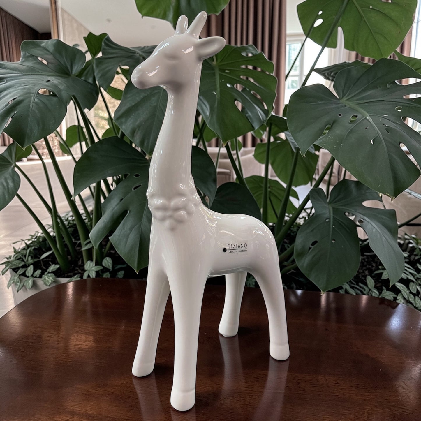 Eine weiße Keramik-Giraffe wird einzeln auf einem Holztisch vor großen grünen Pflanzen im Wohnraum in Szene gesetzt.