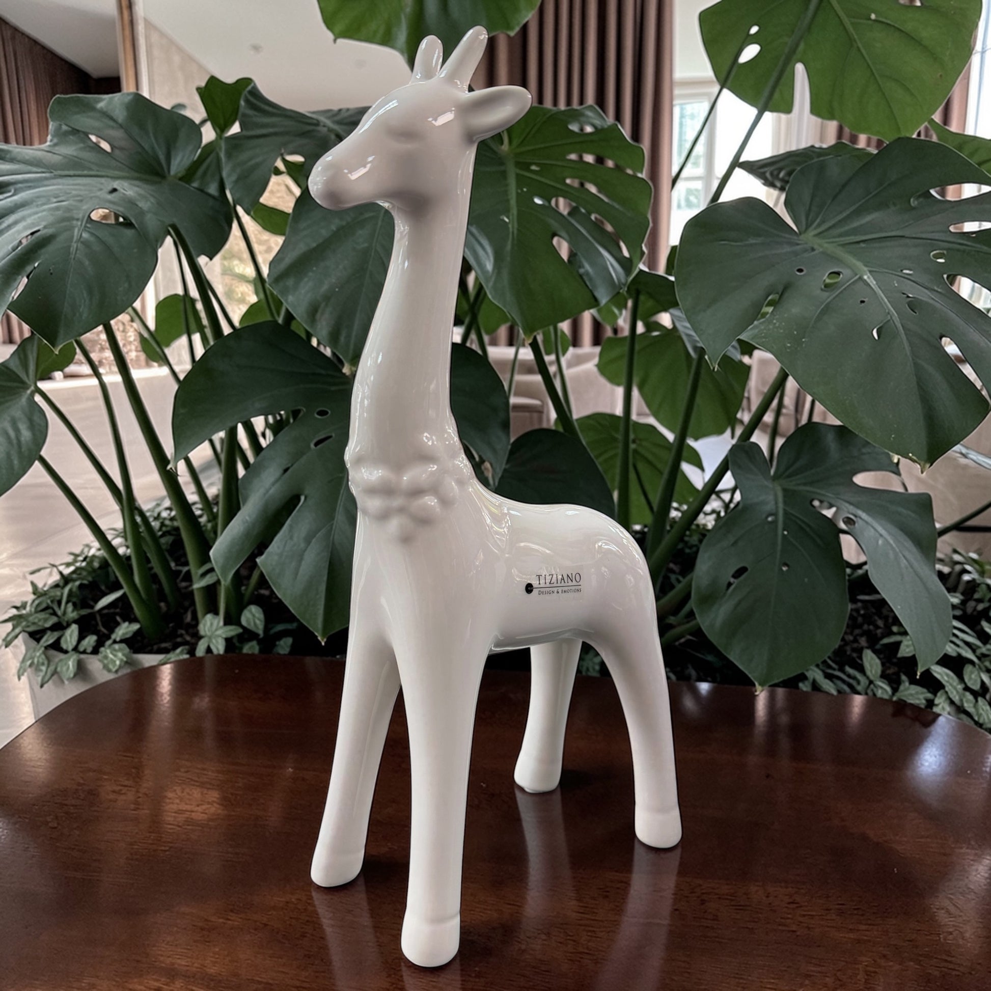 Eine weiße Keramik-Giraffe wird einzeln auf einem Holztisch vor großen grünen Pflanzen im Wohnraum in Szene gesetzt.