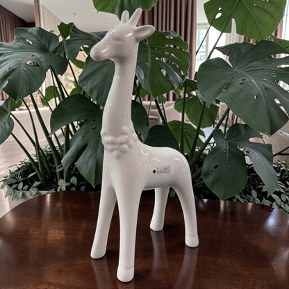 Eine weiße Keramik-Giraffe wird einzeln auf einem Holztisch vor großen grünen Pflanzen im Wohnraum in Szene gesetzt.