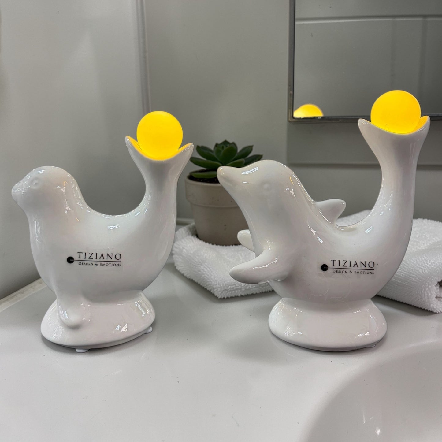 Zwei weiße Keramikfiguren von Tiziano – Robbe und Delfin – mit leuchtender LED-Kugel setzen stilvolle Lichtakzente im Badezimmer.