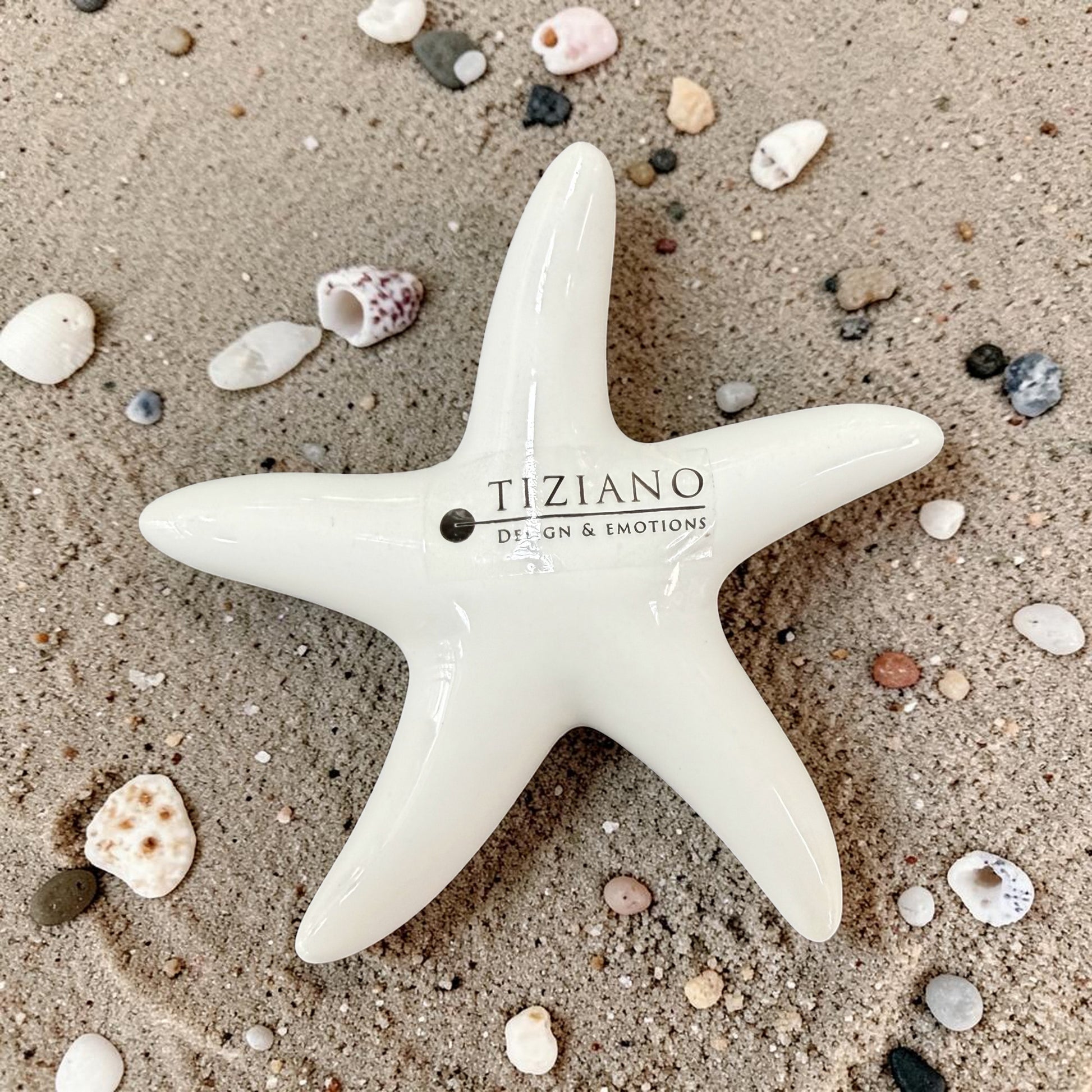 Weiße Seestern-Dekoration mit glänzender Oberfläche liegt auf Sand zwischen kleinen Muscheln und Steinen.