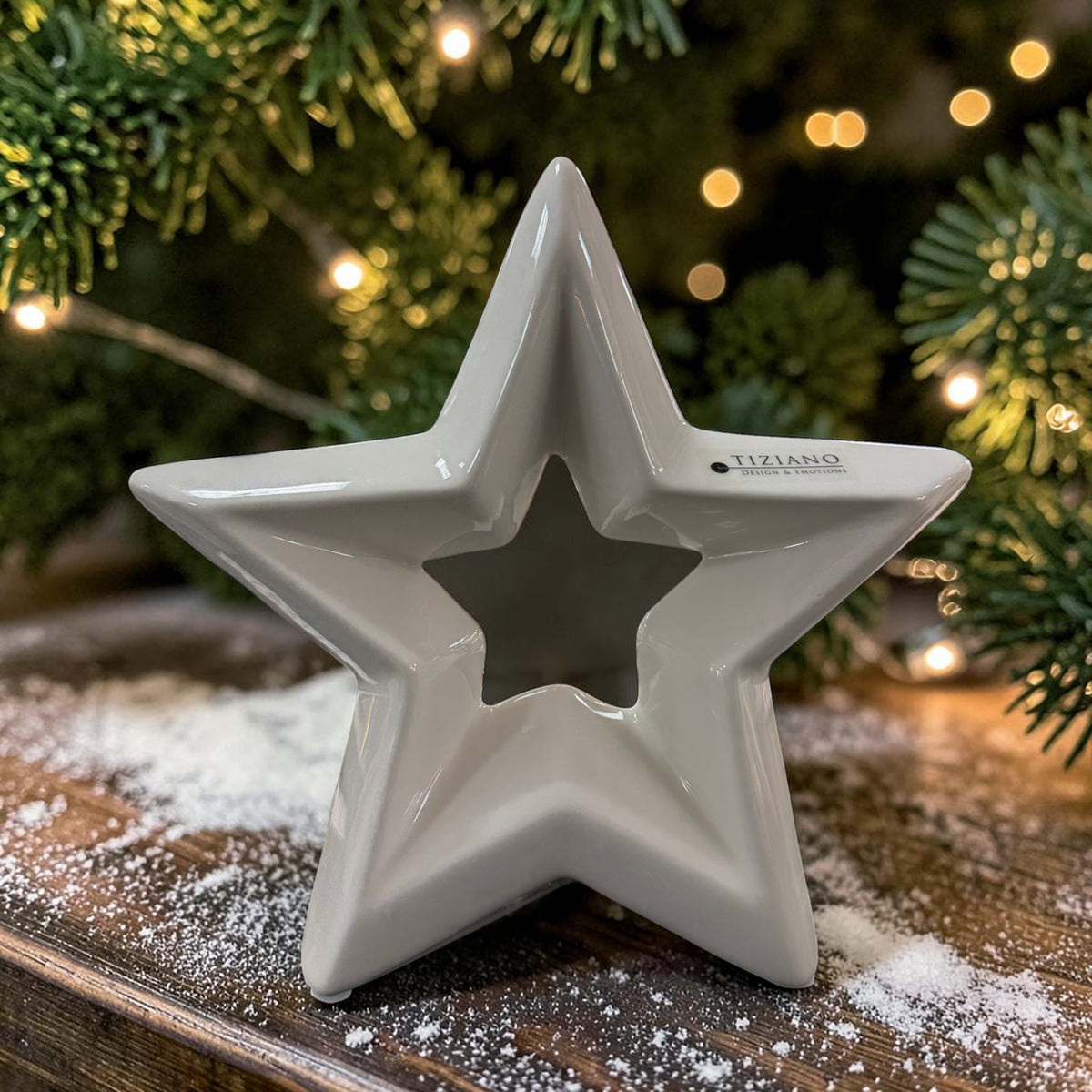 Cremeweißer Tiziano Keramik Stern mit sternförmiger Öffnung auf einem Holztisch zwischen Tannengrün und warmweißer Lichterkette.