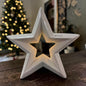 Cremeweißer Tiziano Keramik Stern mit sternförmiger Öffnung und warmweißer Beleuchtung vor einem geschmückten Weihnachtsbaum.