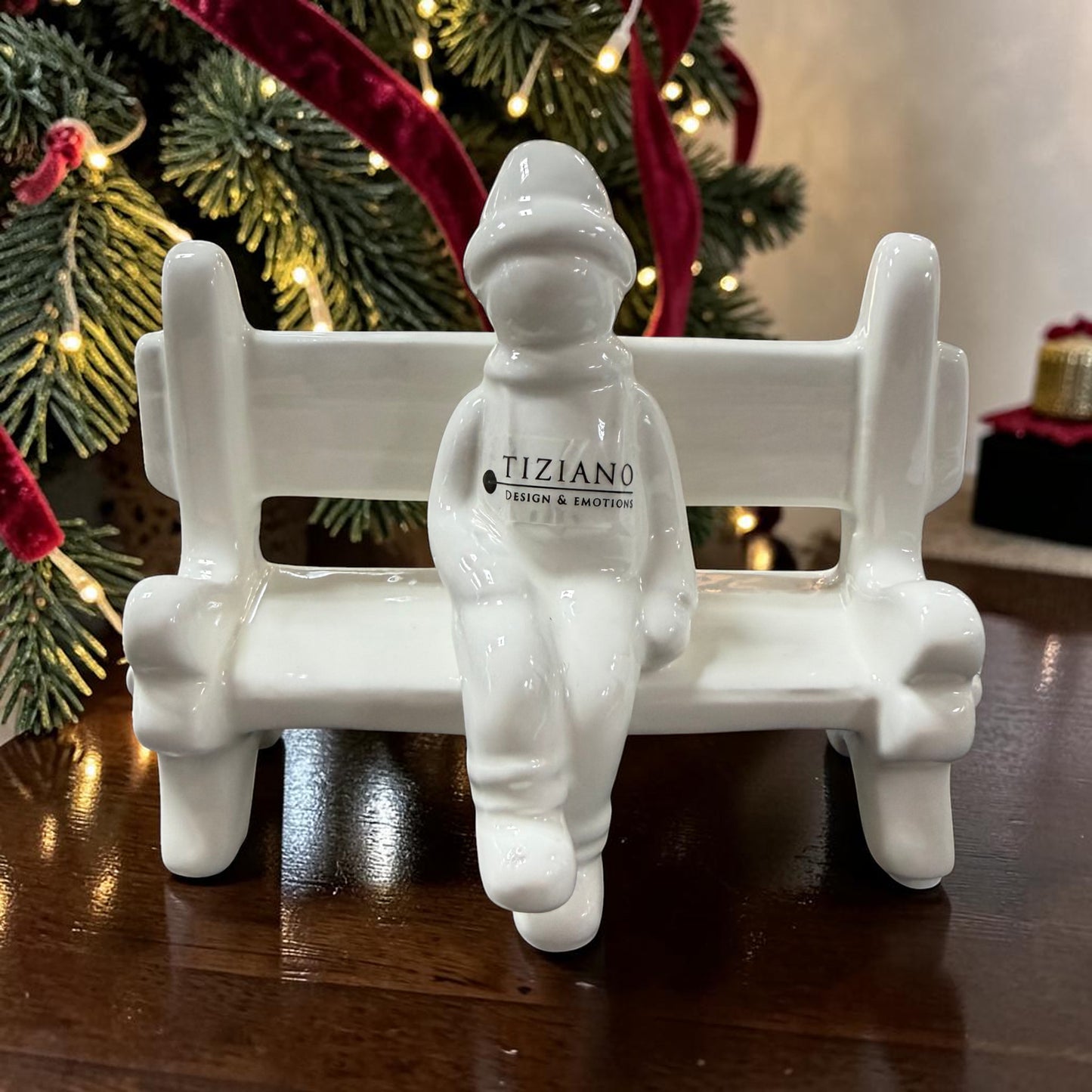 Cremeweiße Tiziano Keramikbank mit einer sitzenden Winterfigur vor einem Weihnachtsbaum mit warm leuchtender Lichterkette.
