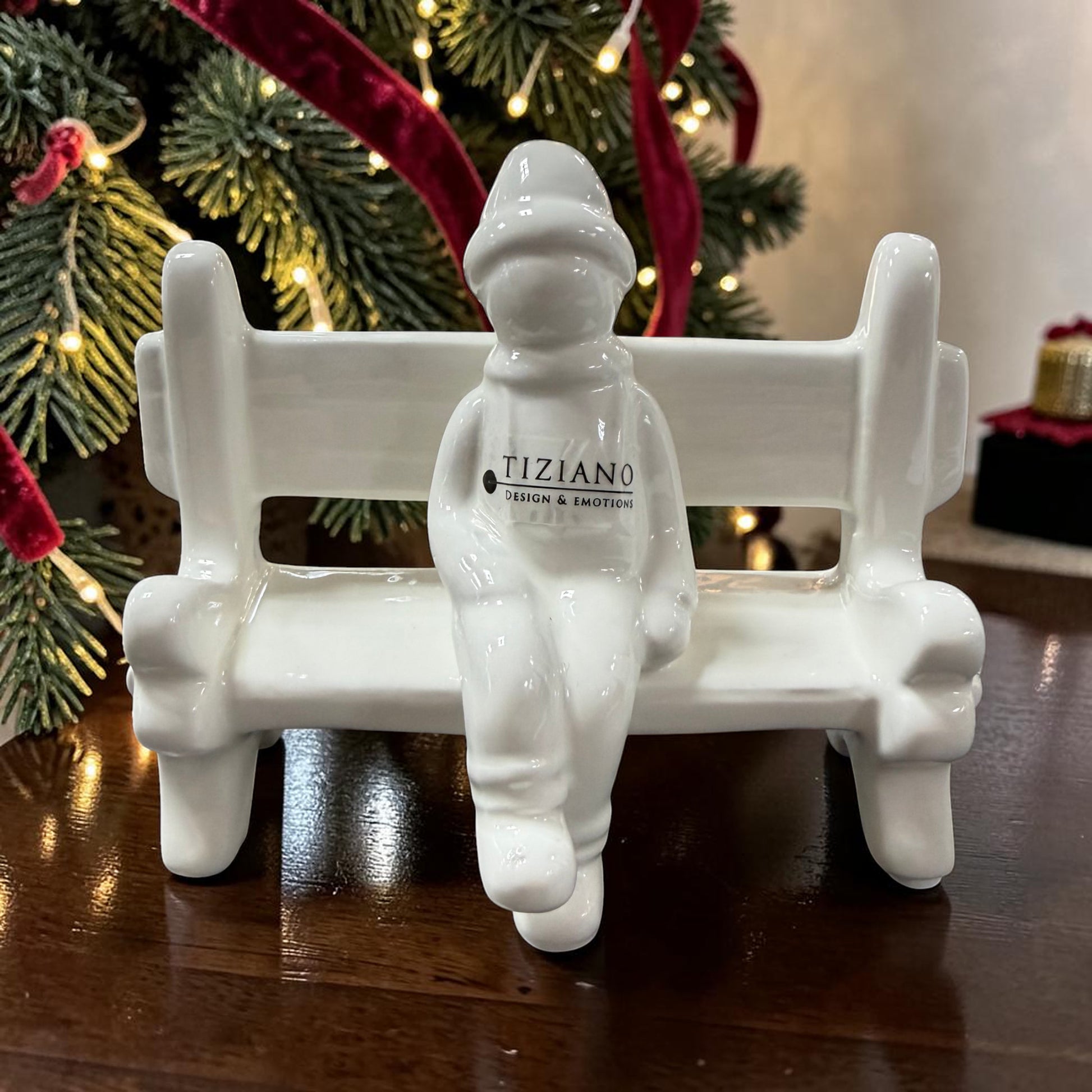 Cremeweiße Tiziano Keramikbank mit einer sitzenden Winterfigur vor einem Weihnachtsbaum mit warm leuchtender Lichterkette.