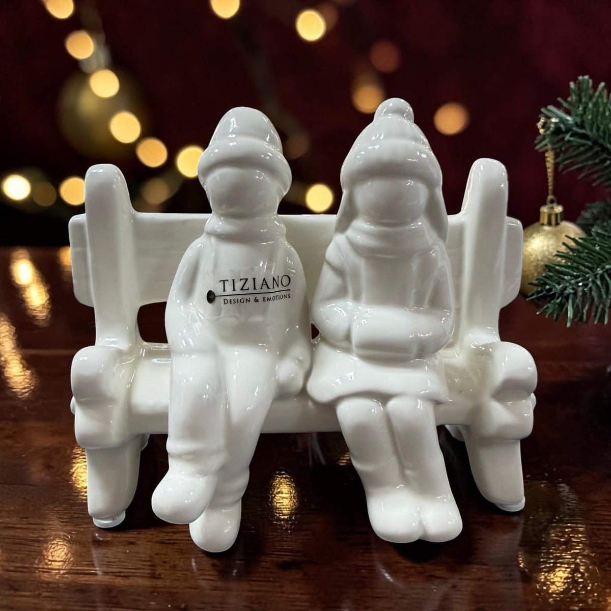 Cremeweiße Tiziano Keramikbank mit zwei sitzenden Winterfiguren vor warmen Lichtern und Tannengrün.