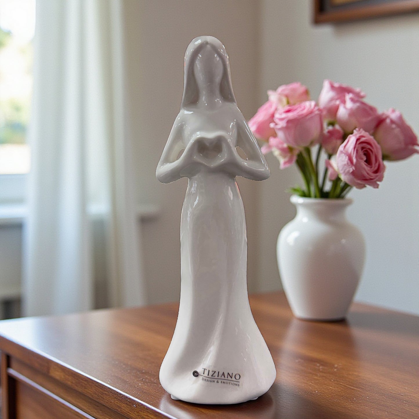 Weiße Keramik-Frauenfigur mit geformtem Herz in den Händen auf einem Holztisch vor Vase mit rosa Blumen.