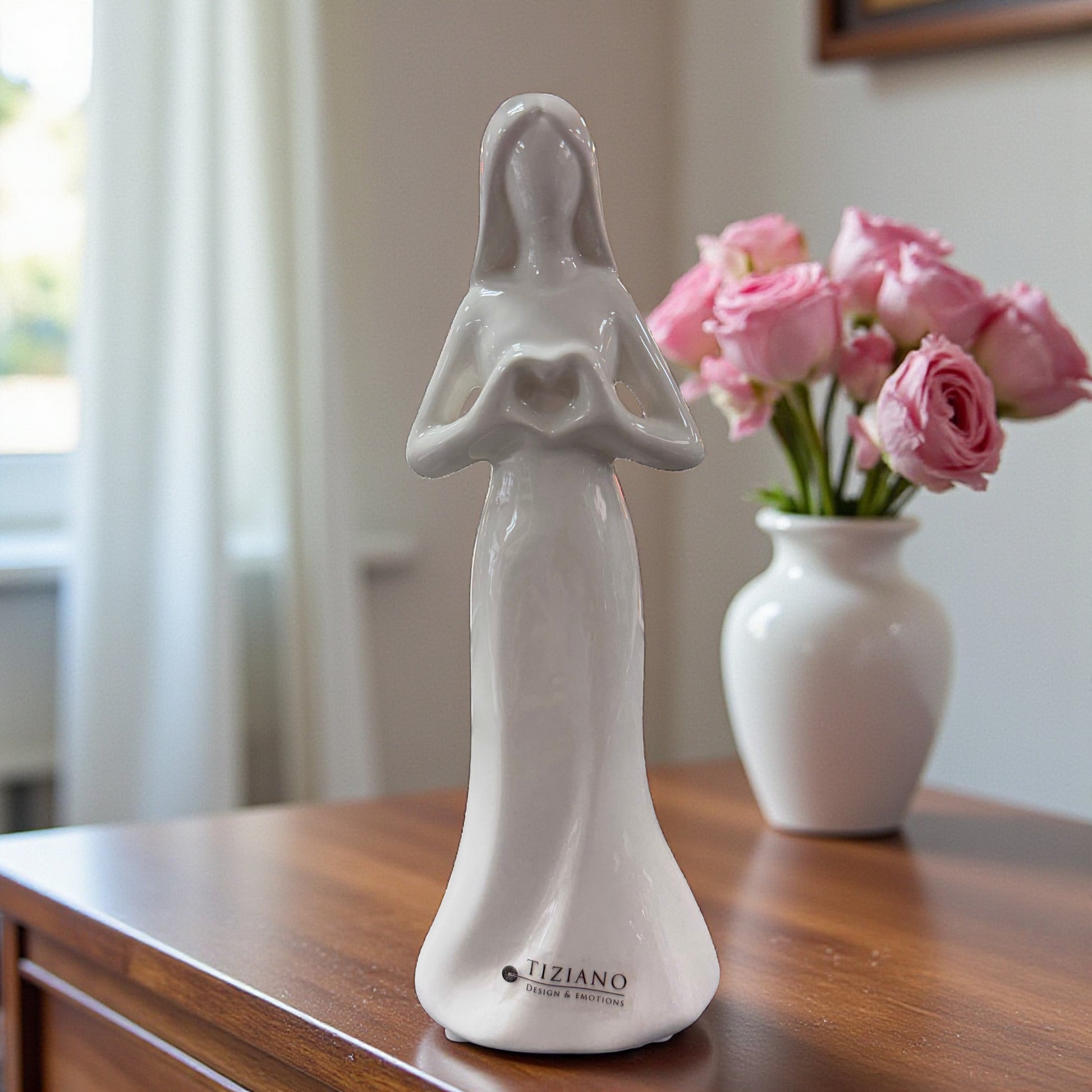 Weiße Keramik-Frauenfigur mit geformtem Herz in den Händen auf einem Holztisch vor Vase mit rosa Blumen.