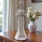 Weiße Keramik-Frauenfigur mit Herz in den Händen auf einem Holztisch neben Vase mit weißen Blumen.