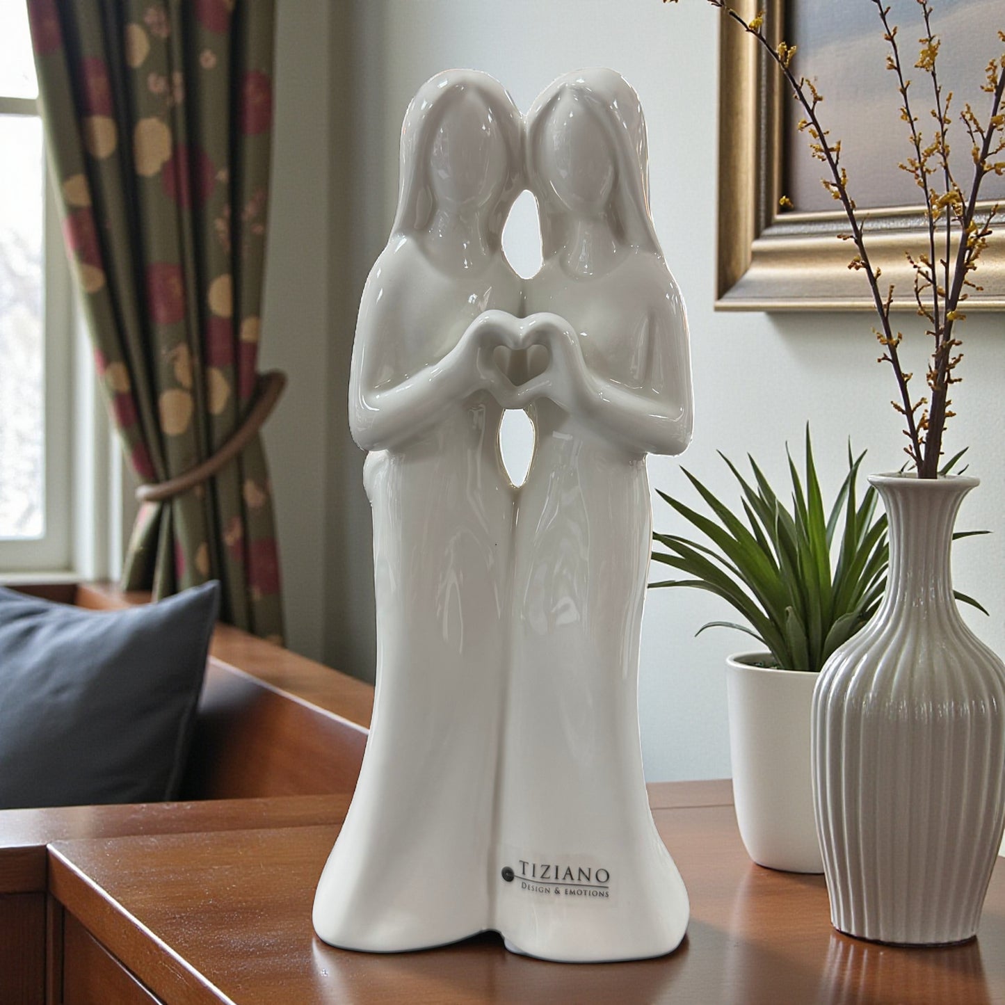 Weiße Keramikfigur eines Frauenpaares mit Herzform aus den Händen auf einem Möbelstück neben Pflanze und Vase