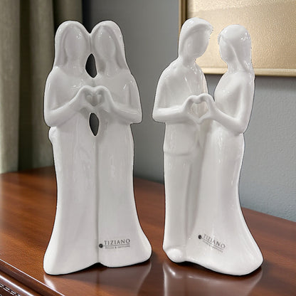 Zwei weiße Keramikfigurenpaare mit Herzform aus den Händen auf einer Holzoberfläche im Innenraum"