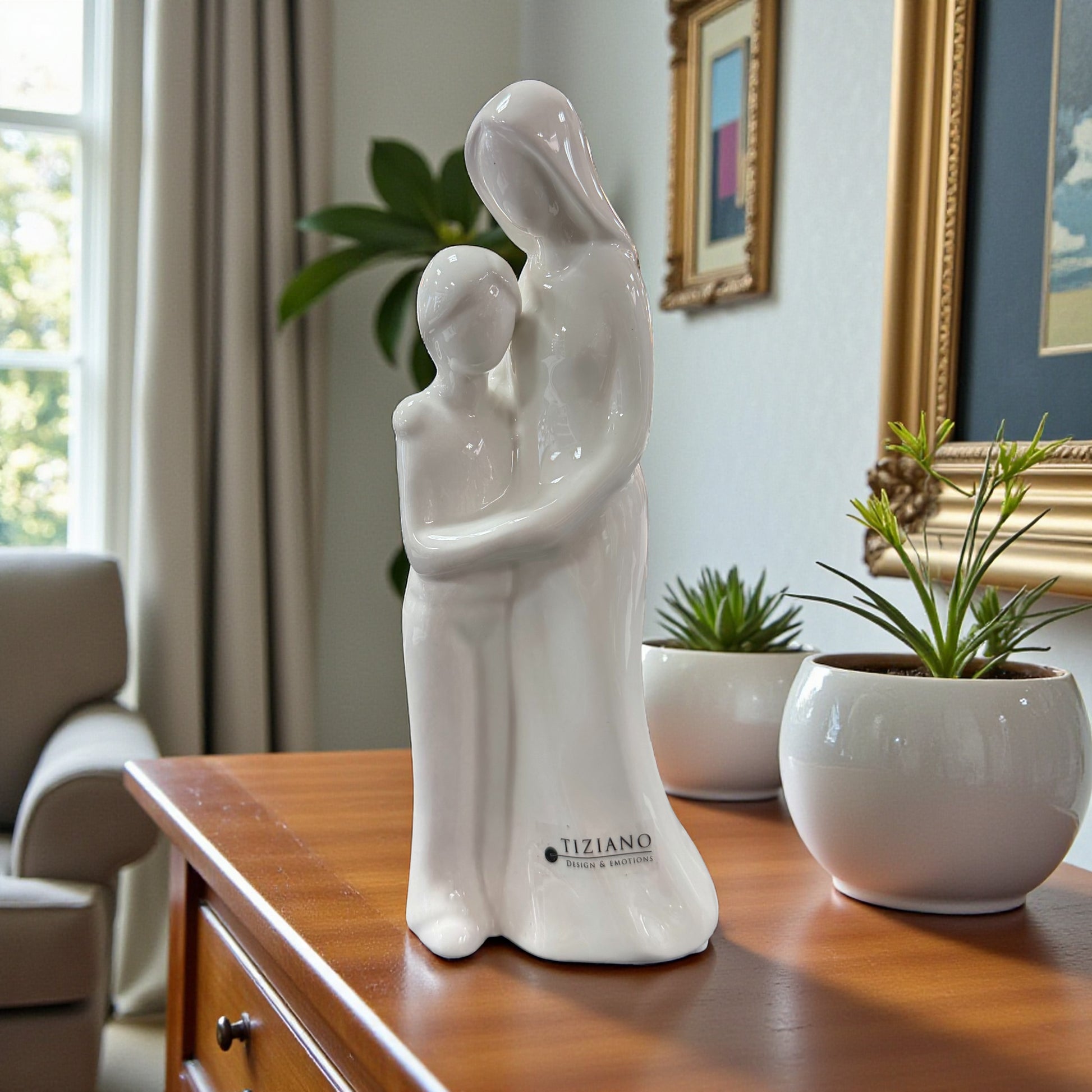 Weiße Keramikfigur mit stilisiertem Mutter-Sohn-Motiv auf einem Möbelstück neben Zimmerpflanzen platziert.