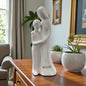 Weiße Keramikfigur mit stilisiertem Mutter-Sohn-Motiv auf einem Möbelstück neben Zimmerpflanzen platziert.