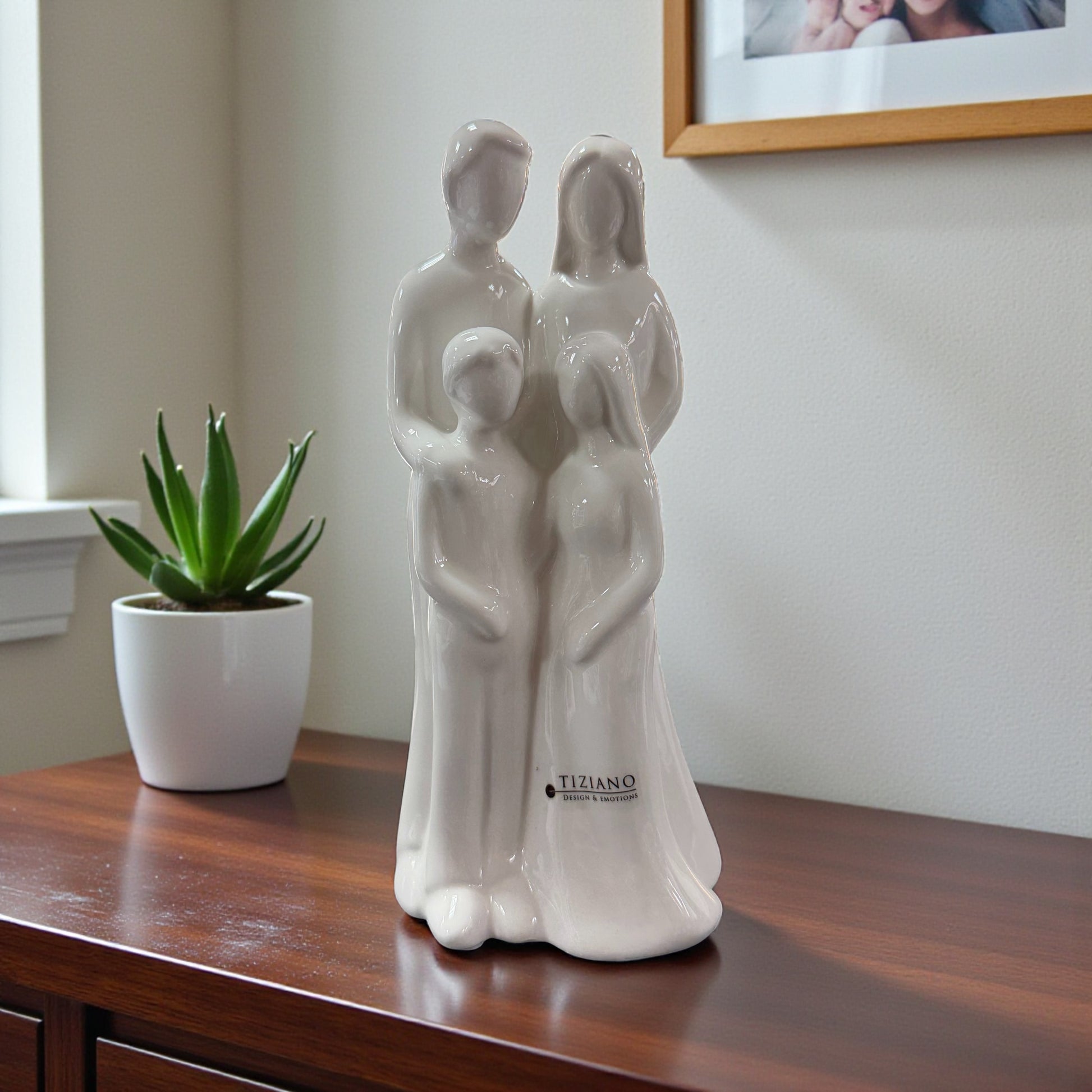 Weiße Keramikfigur mit stilisierter vierköpfiger Familie steht auf einem Möbelstück neben einer Zimmerpflanze.