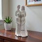 Weiße Keramikfigur mit stilisierter vierköpfiger Familie steht auf einem Möbelstück neben einer Zimmerpflanze.