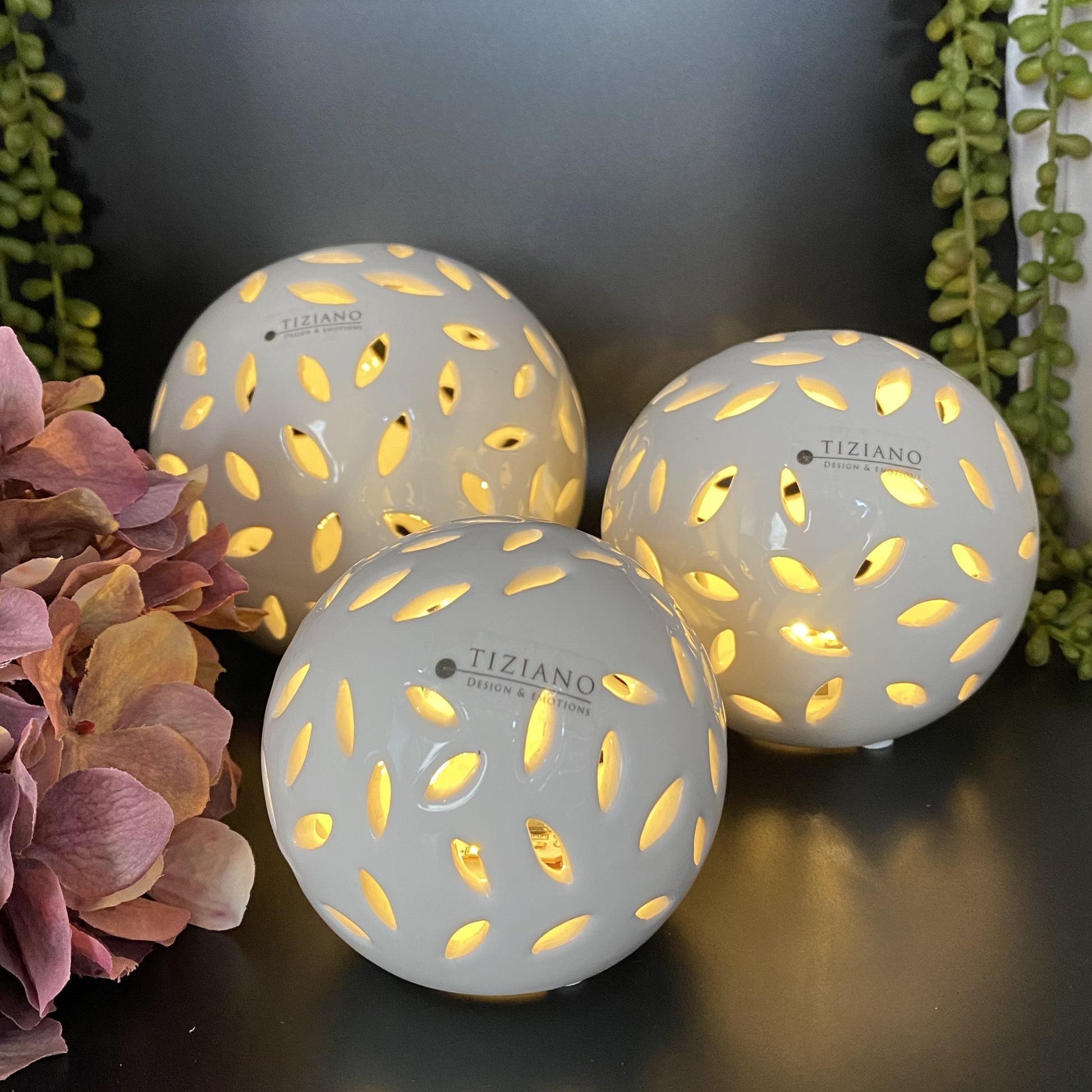 Drei Dekokugeln aus creme-weißer Keramik, die von innen mit einem LED Licht beleuchtet sind. Warmes Licht strahlt aus ovalförmigen Aussparungen. Die Kugeln sind unterschiedlich groß und im Ambiente platziert.