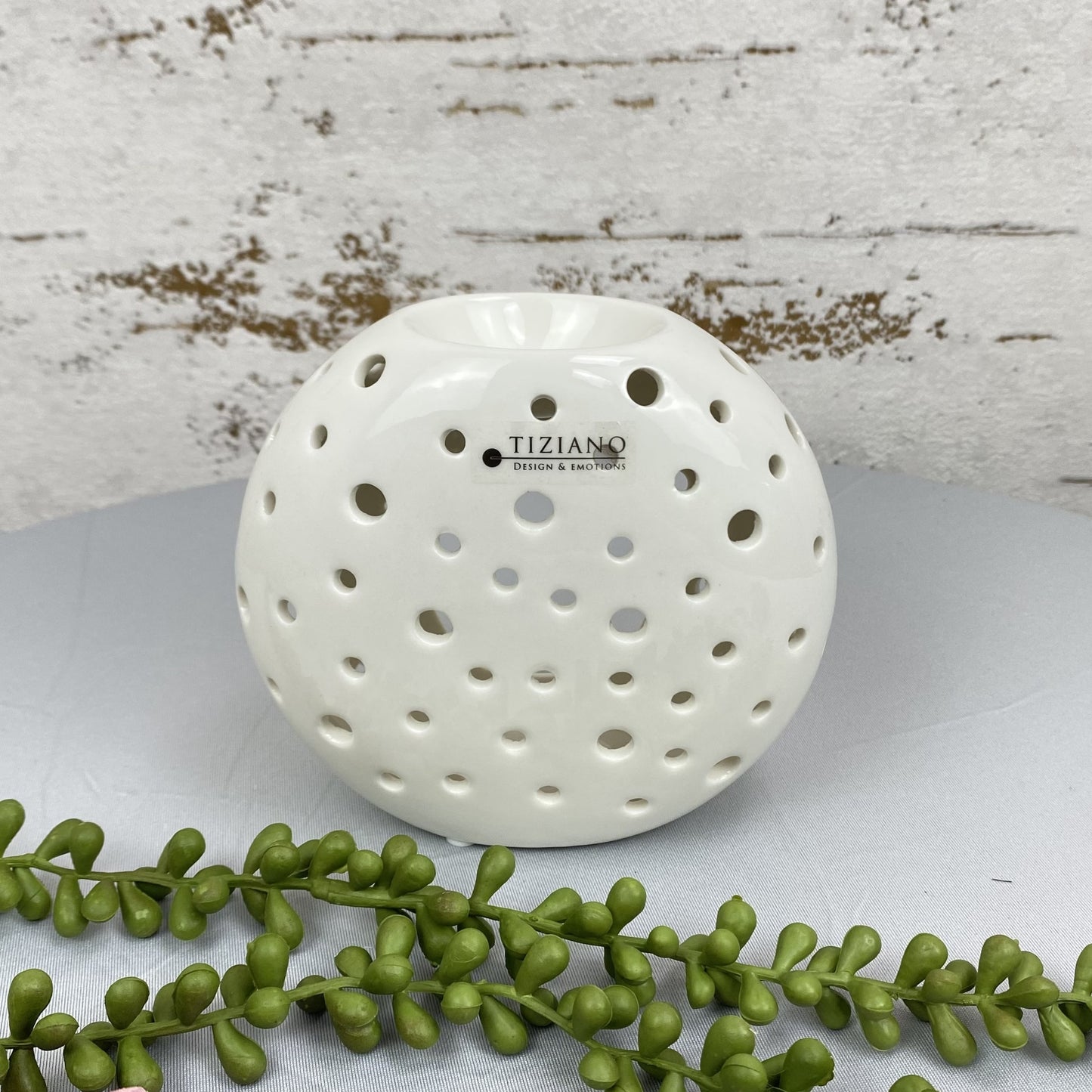 Eine runde Duftlampe für Öl aus creme-weißer Keramik mit runden Aussparungen ist 13cm groß und im AMbiente platziert. 