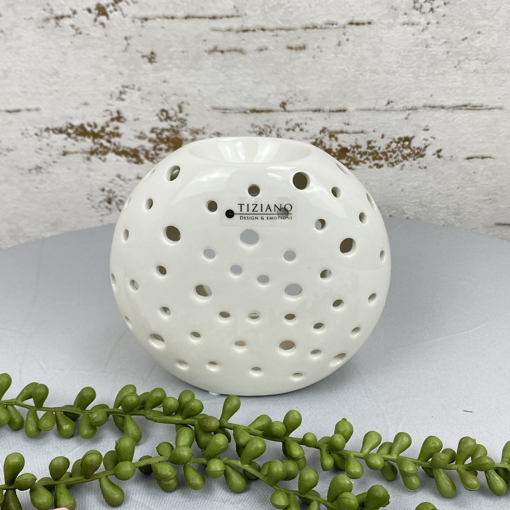 Eine runde Duftlampe für Öl aus creme-weißer Keramik mit runden Aussparungen ist 13cm groß und im AMbiente platziert. 