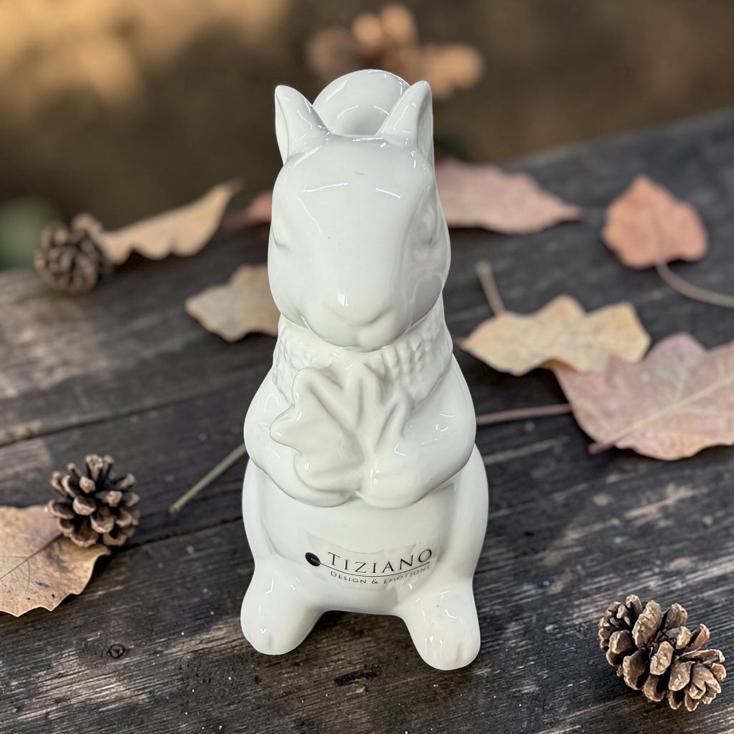 Weißes Tiziano Keramik-Eichhörnchen Robert mit einem Blatt zwischen Herbstlaub und Tannenzapfen.