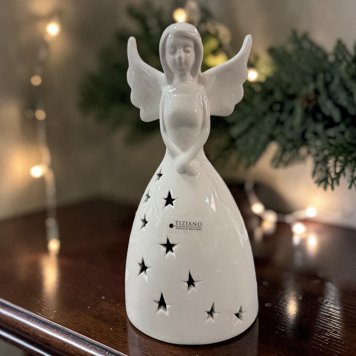 Cremeweißer Tiziano Keramik-Engel mit sternförmigen Ausschnitten unbeleuchtet auf einem dunklen Holztisch mit Tannengrün und warmen Lichtpunkten im Hintergrund.