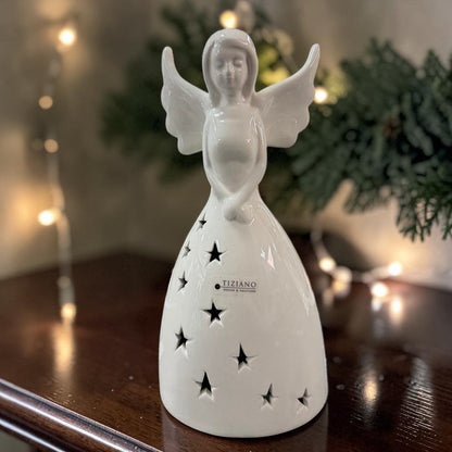 Cremeweißer Tiziano Keramik-Engel mit sternförmigen Ausschnitten unbeleuchtet auf einem dunklen Holztisch mit Tannengrün und warmen Lichtpunkten im Hintergrund.