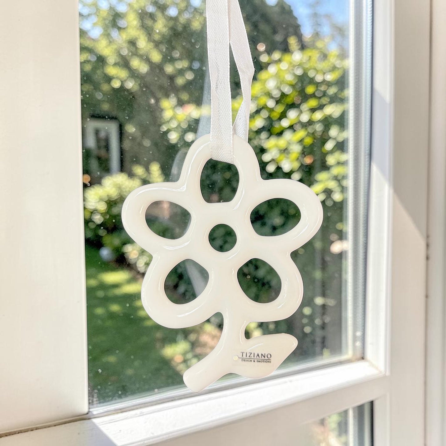 Großer cremeweißer Tiziano Keramik-Fensterhänger in Blumenform mit weißem Band an einem Fenster vor grünem Hintergrund.