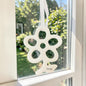 Großer cremeweißer Tiziano Keramik-Fensterhänger in Blumenform mit weißem Band an einem Fenster vor grünem Hintergrund.