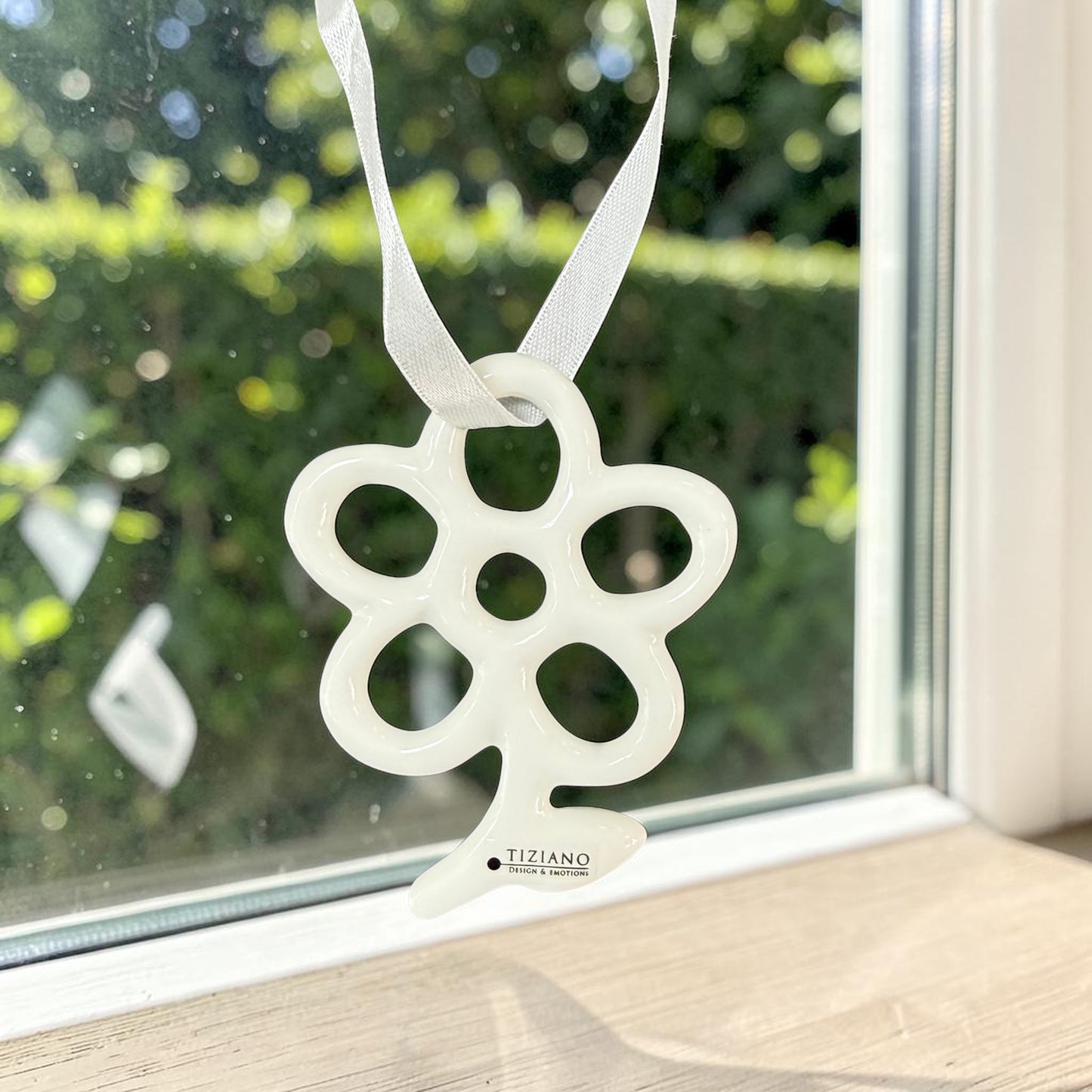 Kleiner cremeweißer Tiziano Keramik-Fensterhänger in Blumenform mit weißem Band vor einem Fenster mit grünem Garten im Hintergrund.