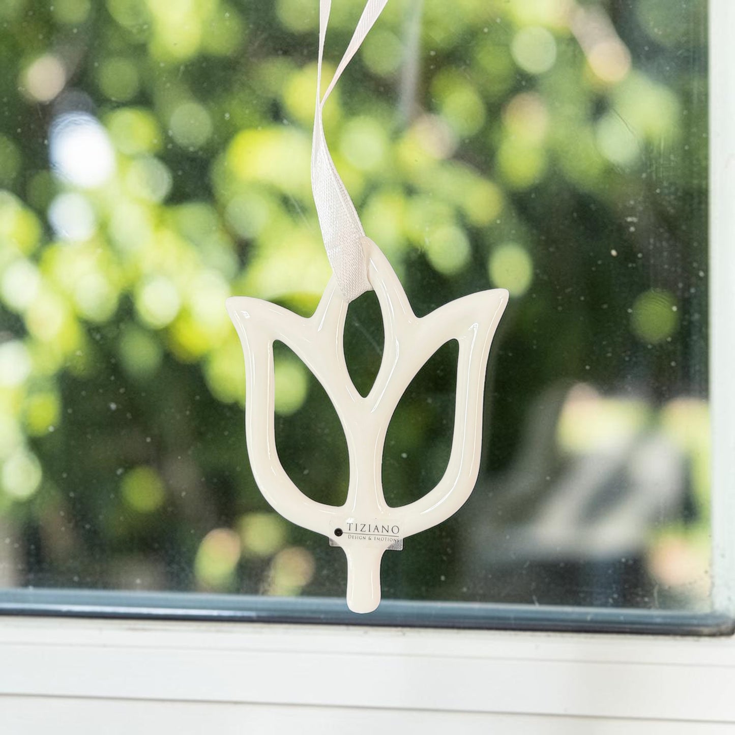 Cremeweiße Tiziano Keramik-Tulpe mit offenem Design und weißem Aufhängeband vor einem Fenster mit grünem Hintergrund.