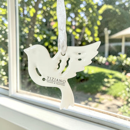 Cremeweißer Tiziano Keramik-Fensterhänger in Vogelform mit weißem Band vor einem Fenster mit grünem Garten im Hintergrund.