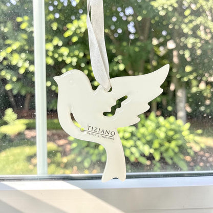 Mittelgroßer cremeweißer Tiziano Keramik-Fensterhänger in Vogelform mit weißem Band vor einem Fenster mit grünem Hintergrund.
