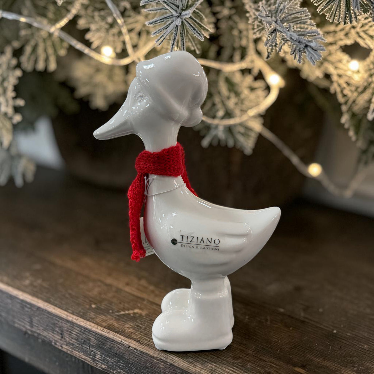 Cremeweiße Tiziano Keramikgans Pato mit rotem Schal auf einem Holztisch unter beschneiten Zweigen und warmer Weihnachtsbeleuchtung.