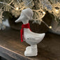 Cremeweiße Tiziano Keramikgans Pato mit rotem Schal auf einem Holztisch unter beschneiten Zweigen und warmer Weihnachtsbeleuchtung.