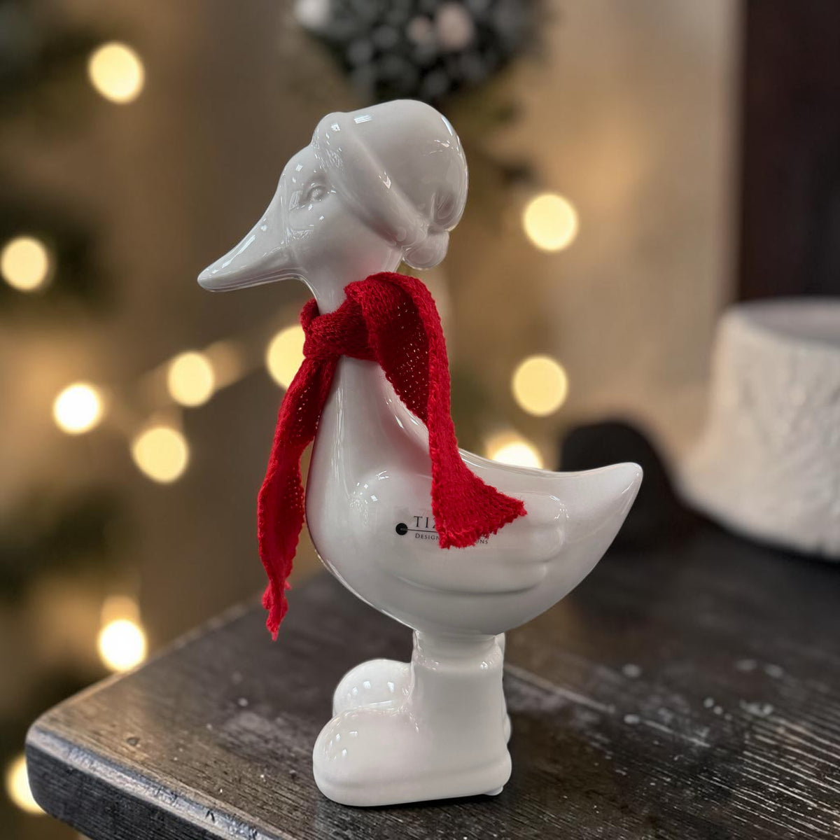 Cremeweiße Tiziano Keramikgans Pato mit rotem Schal auf einem dunklen Holztisch vor unscharfer Weihnachtsbeleuchtung.