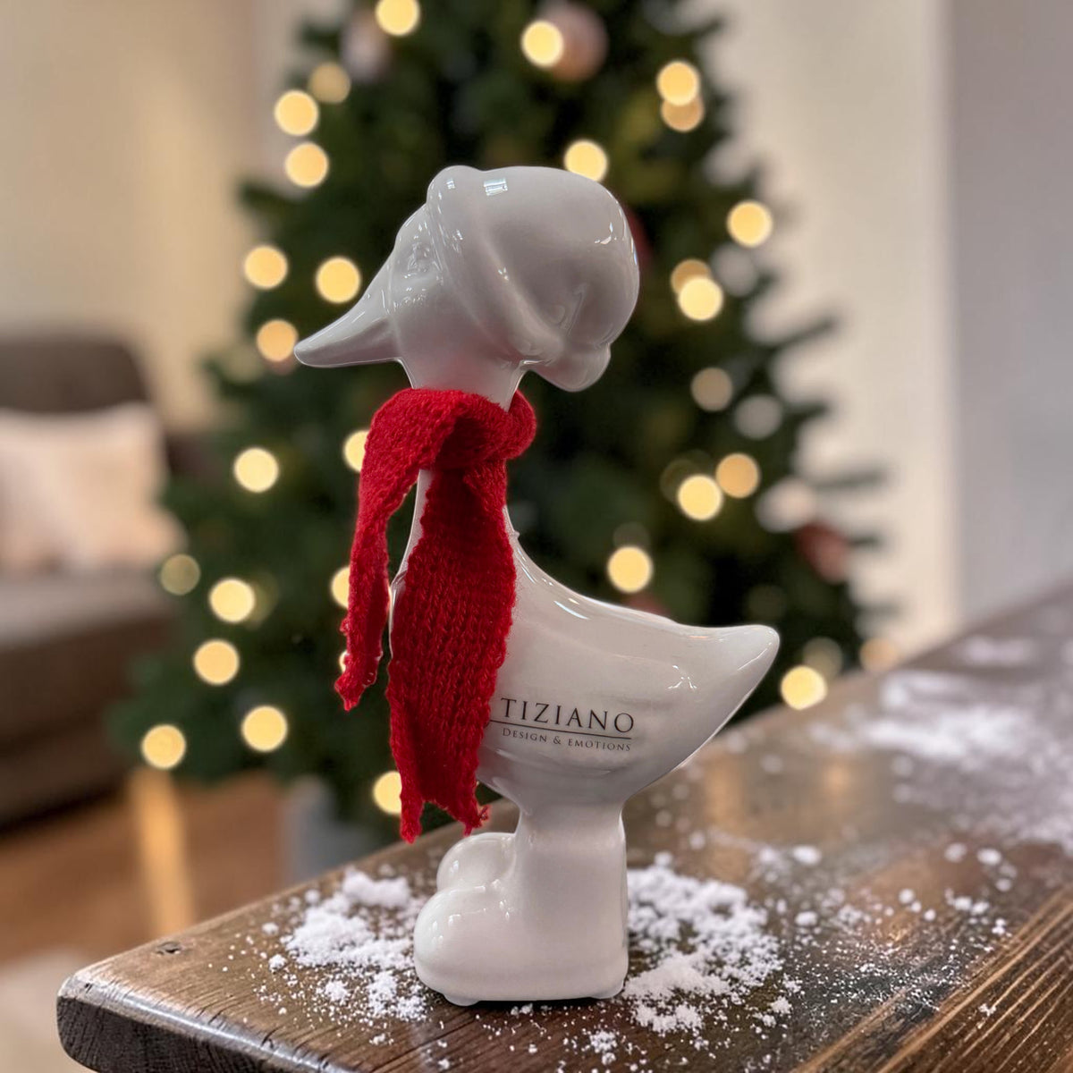 Cremeweiße Tiziano Keramikgans Pato mit rotem Schal auf einem Holztisch mit Kunstschnee vor einem beleuchteten Weihnachtsbaum.