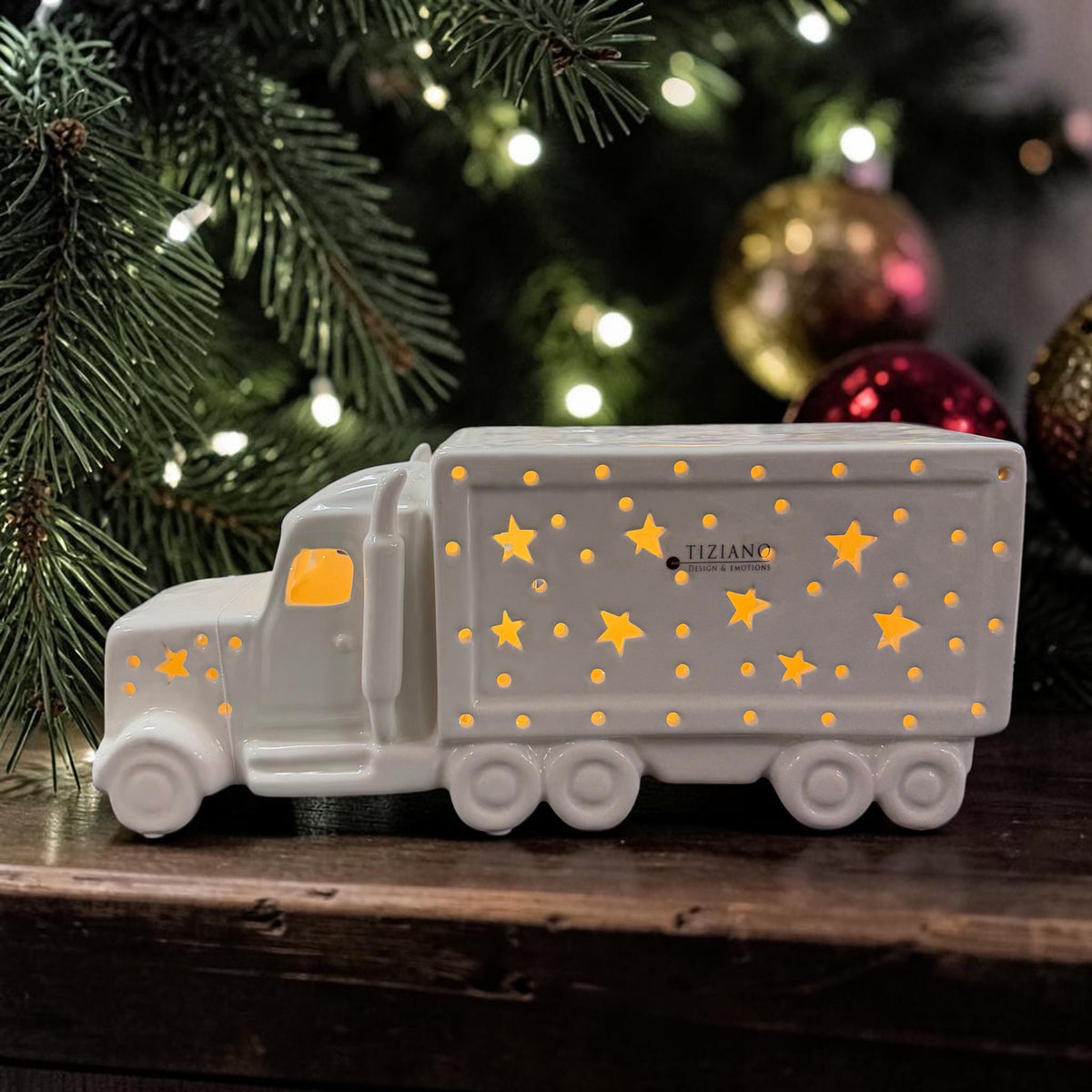 Cremeweißer Tiziano Keramik LED-LKW mit warm leuchtenden Sternen vor einem Weihnachtsbaum mit Lichterkette sowie goldenen und roten Weihnachtskugeln.