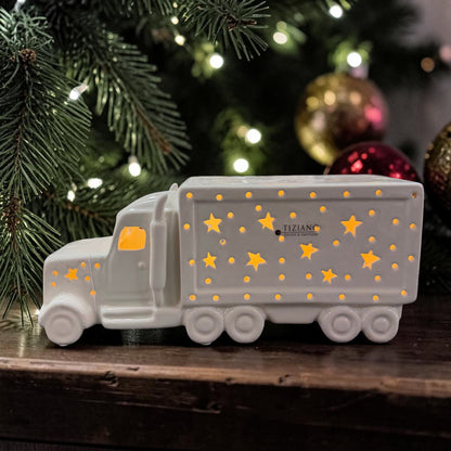 Cremeweißer Tiziano Keramik LED-LKW mit warm leuchtenden Sternen vor einem Weihnachtsbaum mit Lichterkette sowie goldenen und roten Weihnachtskugeln.