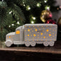 Cremeweißer Tiziano Keramik LED-LKW mit warm leuchtenden Sternen vor einem Weihnachtsbaum mit Lichterkette sowie goldenen und roten Weihnachtskugeln.