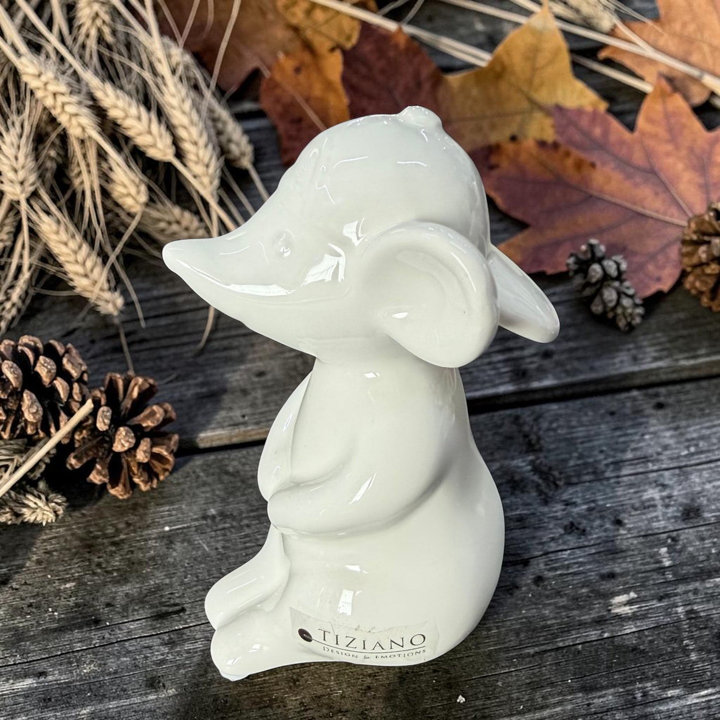 Weiße Tiziano Keramik-Maus mit großen runden Ohren auf einem dunklen Holztisch zwischen buntem Herbstlaub und Getreide.